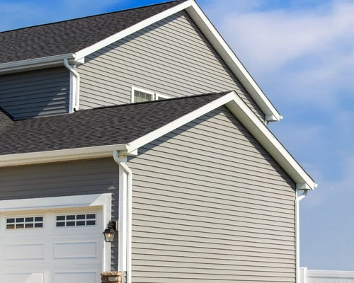 A close-up of a modern house exterior with light gray vinyl siding, white trim, and a white garage door under a blue sky. From Green Built Roofing in Michigan.