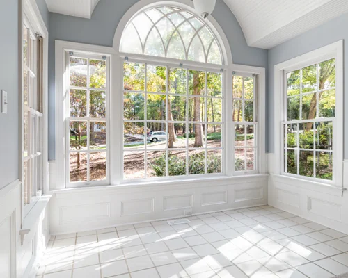 A bright sunroom with large arched and rectangular windows, white tile floor, and light blue-gray walls, overlooking a wooded yard. From Green Built Roofing in Michigan.