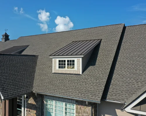 A close-up view of a house roof with gray asphalt shingles and a dormer window with a metal roof under a clear blue sky. From Green Built Roofing in Michigan.